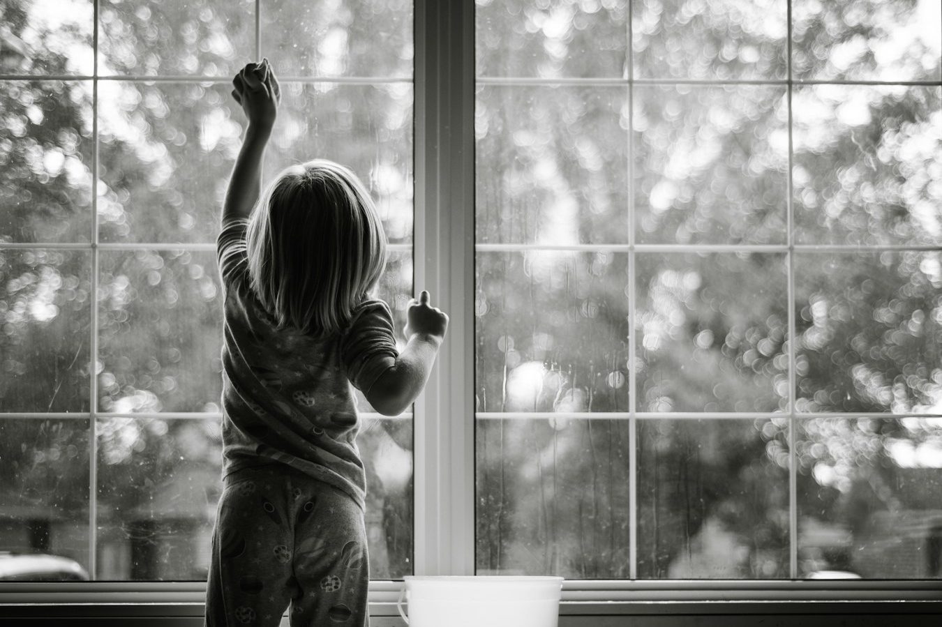 Ein Kind steht am Fenster und schaut nach draußen in einen bewaldeten Garten.